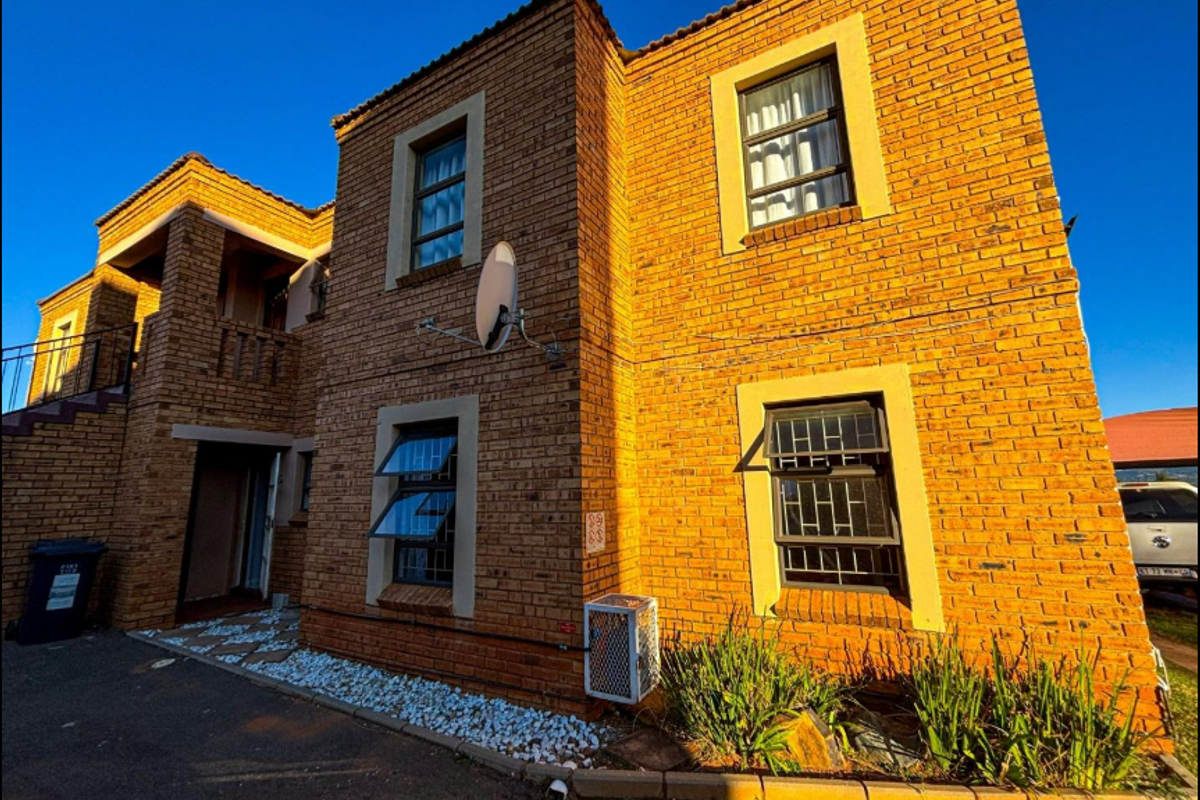A two-story face-brick townhouse exterior featuring top-hung windows with security bars, a paved driveway, and an external air conditioning unit.