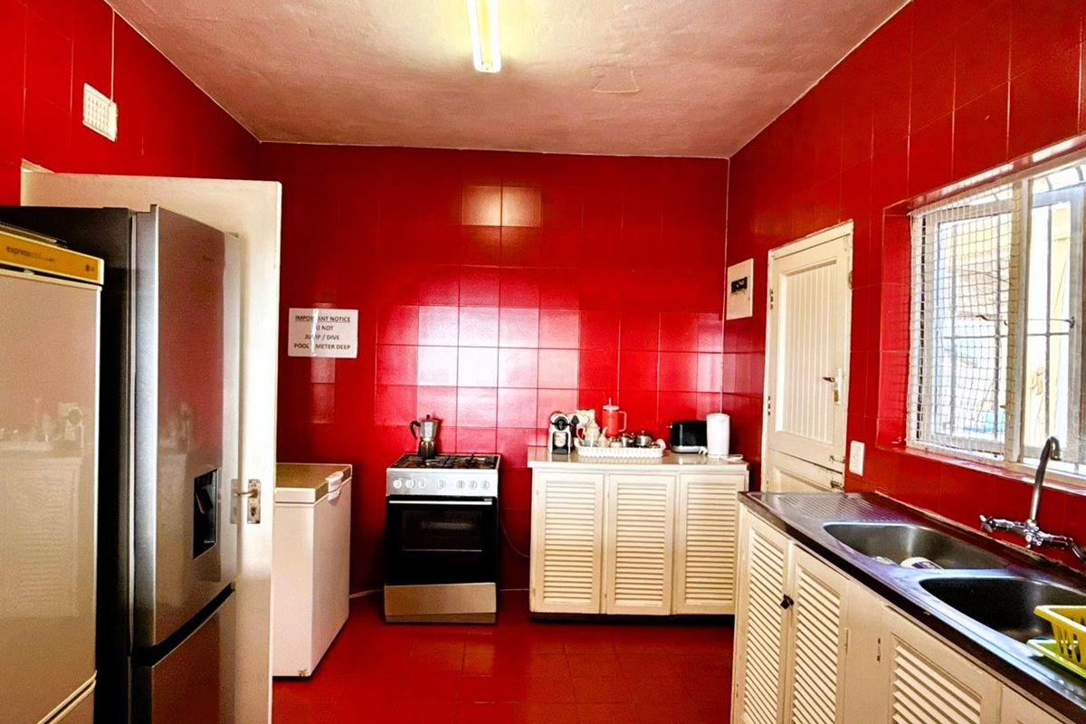 A dated kitchen featuring bold red wall and floor tiles, white louvered cabinetry, a stainless steel sink, and a freestanding gas stove.