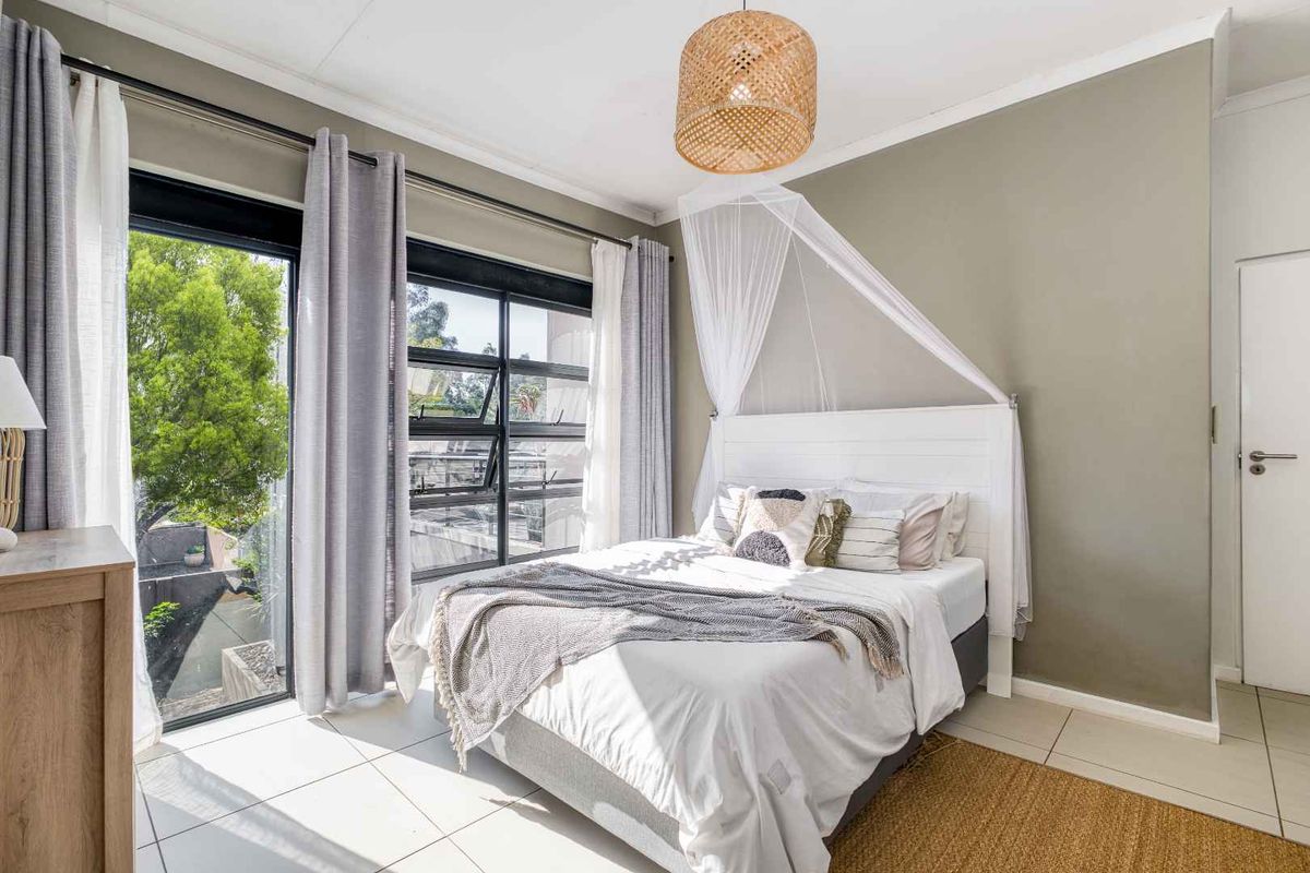 A bright and well-maintained bedroom featuring tiled flooring, neutral wall tones, and large aluminium-framed windows providing natural light and outdoor access.