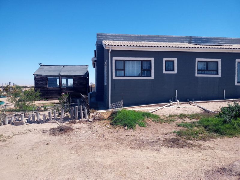 Rear view of the property showing the main dwelling with tiled roof and a separate wooden outbuilding structure set on a sandy plot.