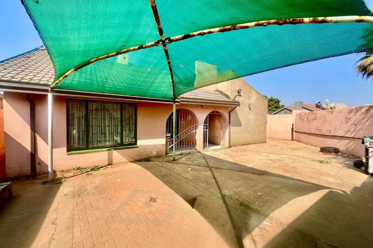 Rear exterior view featuring a paved patio area covered by a green shade sail, with security gates and bars on the building facade.