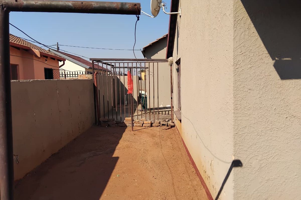 A side exterior view of the property featuring a metal security gate, exposed soil ground, and a satellite dish mounted on the exterior wall.