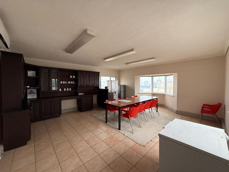 Spacious open-plan area featuring tiled flooring, built-in dark wood cabinetry, and large bay windows providing natural light, suitable for office or breakroom use.