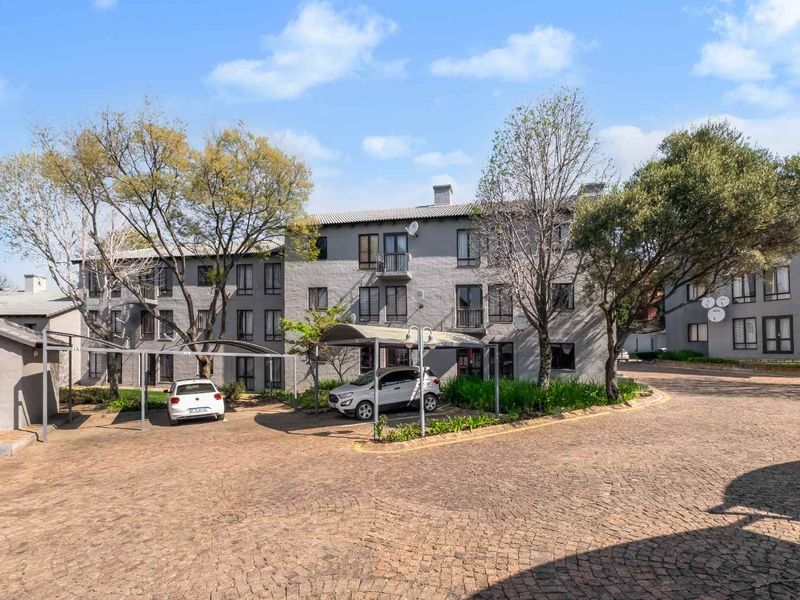 Exterior view of a multi-story apartment complex featuring a paved driveway, covered parking bays, and established trees.