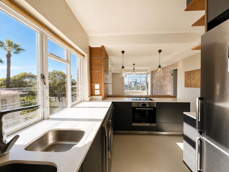 Modern kitchen featuring sleek dark cabinetry, stone countertops, built-in oven, cooktop, and an exposed brick feature wall with abundant natural light.