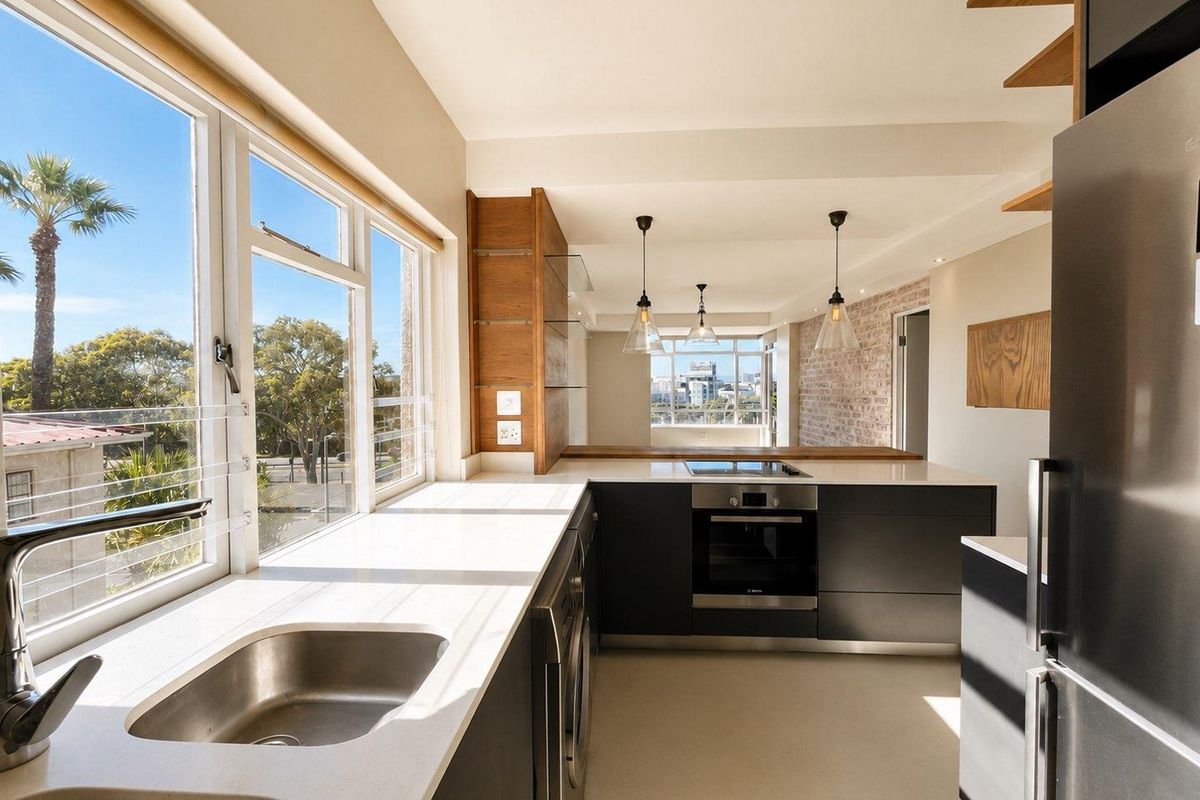 Modern kitchen featuring sleek dark cabinetry, stone countertops, built-in oven, cooktop, and an exposed brick feature wall with abundant natural light.
