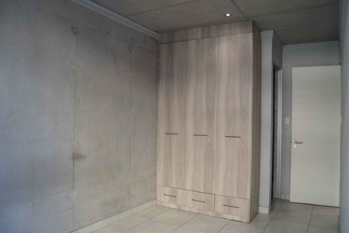 A modern bedroom featuring a built-in wardrobe, tiled flooring, and an exposed concrete ceiling with recessed lighting.