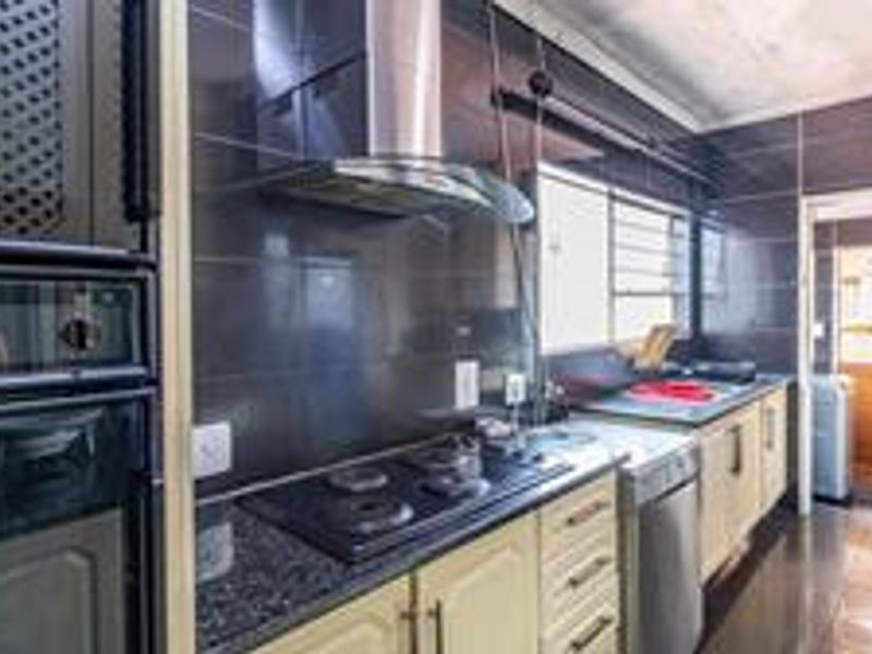 A functional kitchen featuring built-in appliances, dark tiled splashbacks, granite countertops, and ample fitted cabinetry with a view into an adjacent utility area.