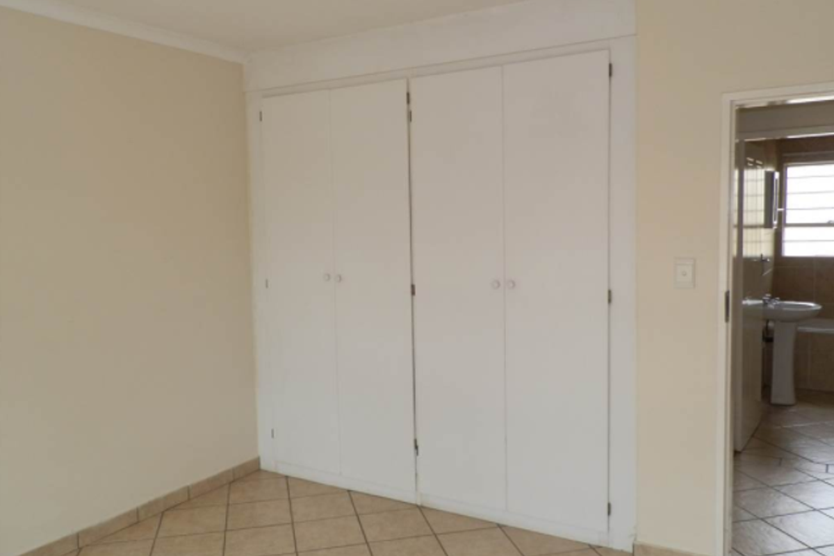 A clean, empty bedroom featuring neutral walls, tiled flooring, and built-in cupboards, with an adjacent view into a tiled bathroom.