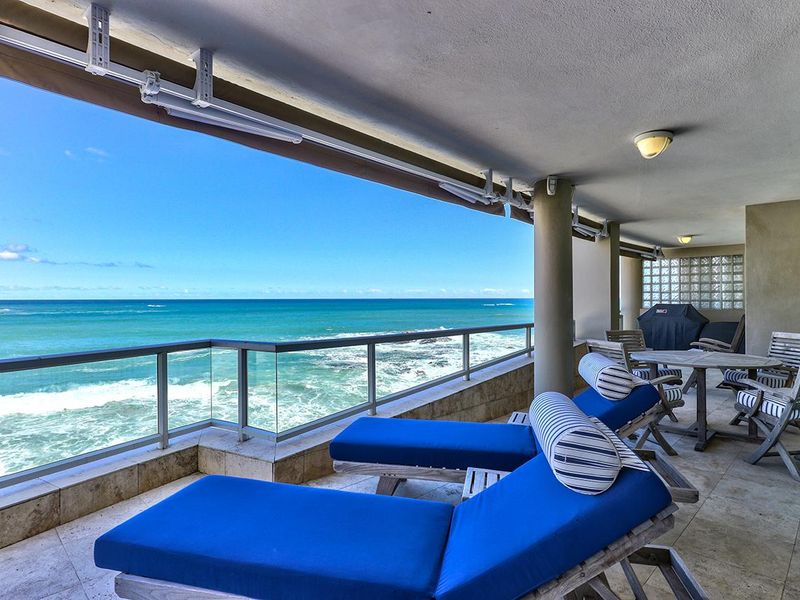 Spacious covered balcony featuring stone flooring, a glass balustrade, and a retractable awning, offering expansive ocean views.