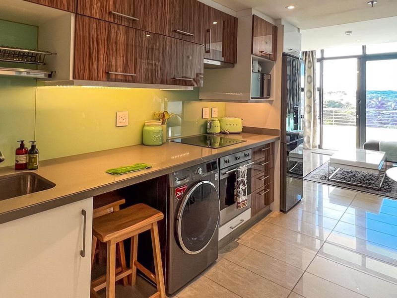 Modern open-plan kitchen featuring sleek cabinetry, integrated appliances, stone countertops, and a glass splashback, flowing seamlessly into the living area with balcony access.