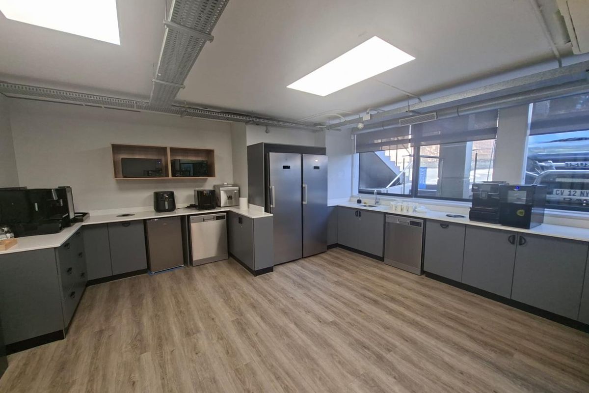 Modern office kitchen area featuring grey cabinetry, integrated appliances, wood-look flooring, and exposed industrial-style ceiling cable trays.