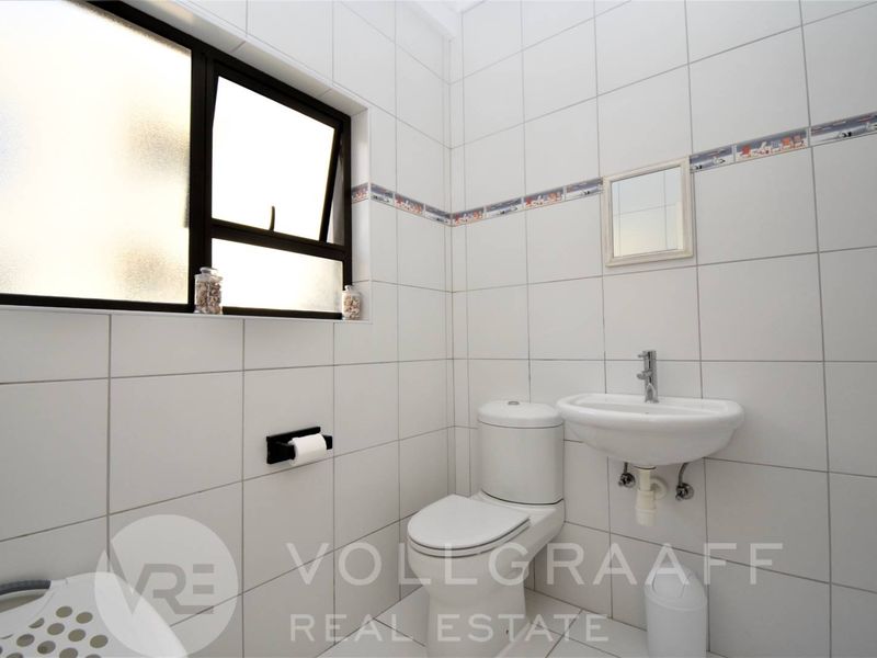 A compact, fully tiled bathroom featuring a white toilet, wall-mounted basin, and a top-hung window for natural light.