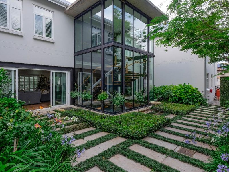 A beautifully landscaped rear garden featuring a striking glass-enclosed staircase structure, paved pathways, and seamless indoor-outdoor flow through sliding glass doors.