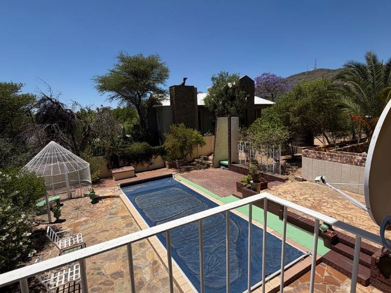 An elevated view of a backyard featuring a swimming pool with a cover, stone paved patio area, and surrounding garden landscaping.