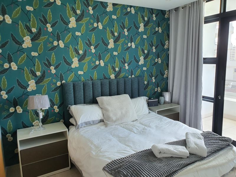 A well-maintained bedroom featuring a bold floral accent wall, neutral carpeting, and direct access to a private balcony through sliding glass doors.