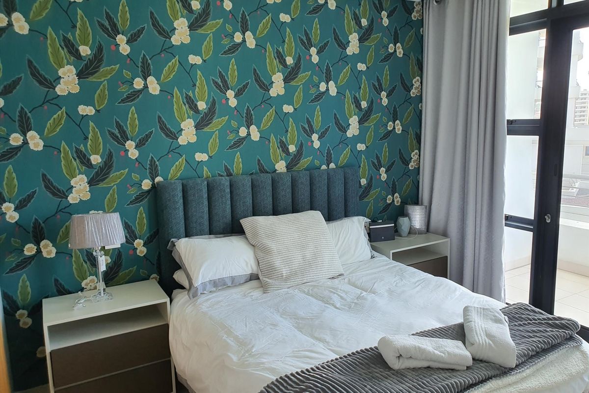 A well-maintained bedroom featuring a bold floral accent wall, neutral carpeting, and direct access to a private balcony through sliding glass doors.