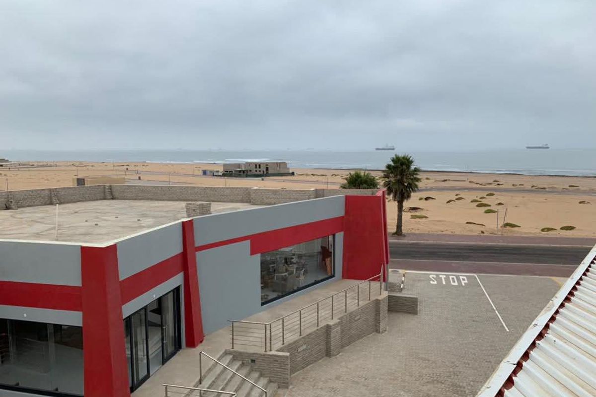 Elevated view of a modern commercial building featuring red and grey facade accents, paved exterior area, and direct proximity to the coastline.
