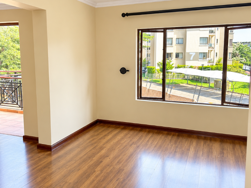 A bright, empty living room featuring warm-toned laminate flooring, crown molding, and a large window providing natural light and views of the complex.