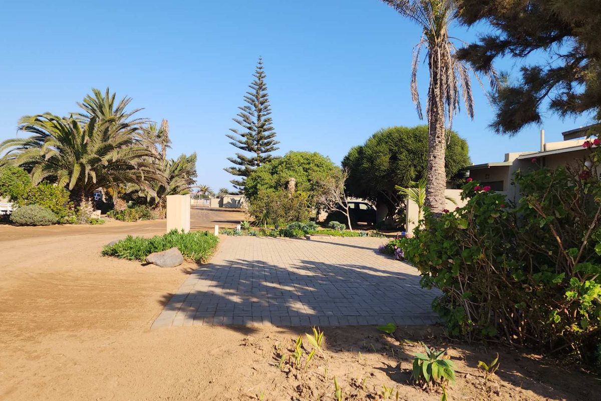 A view of a residential property entrance featuring a paved driveway, established garden landscaping, and mature trees on a sandy plot.