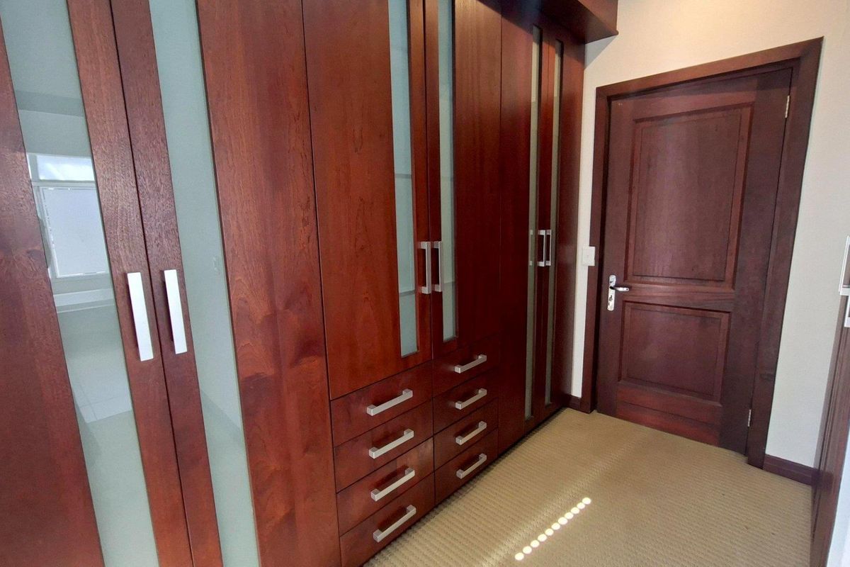A clean bedroom dressing area featuring extensive built-in timber cabinetry with integrated drawers and glass accents, complemented by neutral carpeting.
