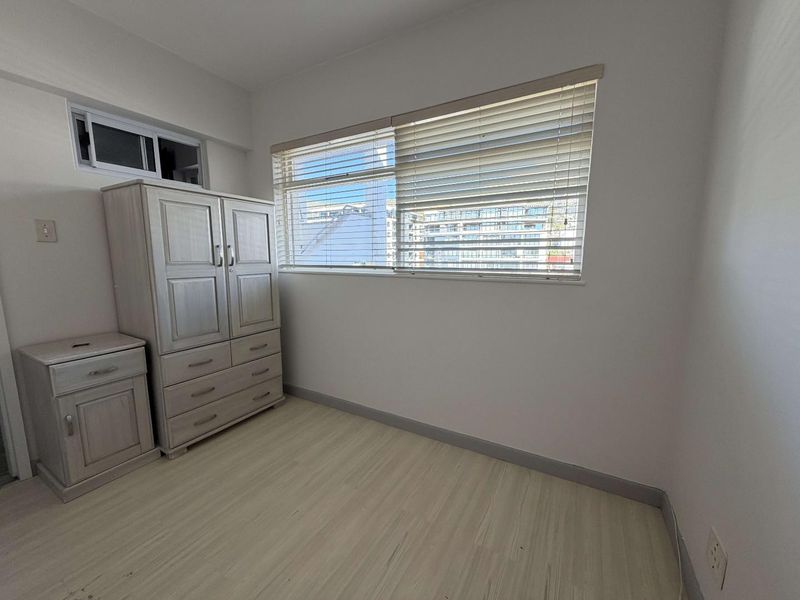 A bedroom featuring light-toned laminate flooring, neutral walls, and venetian blinds on the window, providing a clean and functional living space.