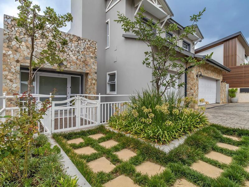 Modern multi-story residential facade featuring stone cladding, a paved driveway, a double garage, and a landscaped front garden with a pedestrian gate.