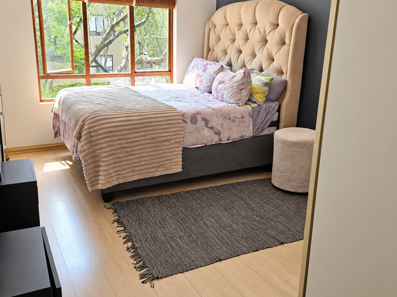A well-lit bedroom featuring light-toned laminate flooring, a large timber-framed window, and a built-in wardrobe.