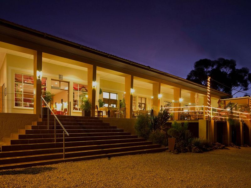 A well-lit hotel exterior at dusk featuring a spacious covered veranda, accessible via a wide concrete staircase with metal railings.