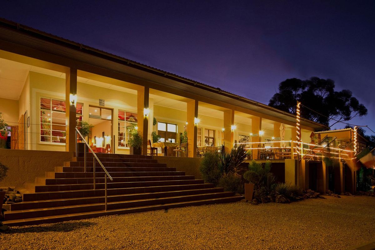 A well-lit hotel exterior at dusk featuring a spacious covered veranda, accessible via a wide concrete staircase with metal railings.