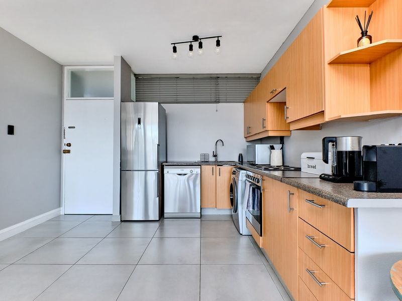 A functional kitchen featuring light wood-finish cabinetry, stone-look countertops, tiled flooring, and integrated appliances, offering a clean and practical workspace.