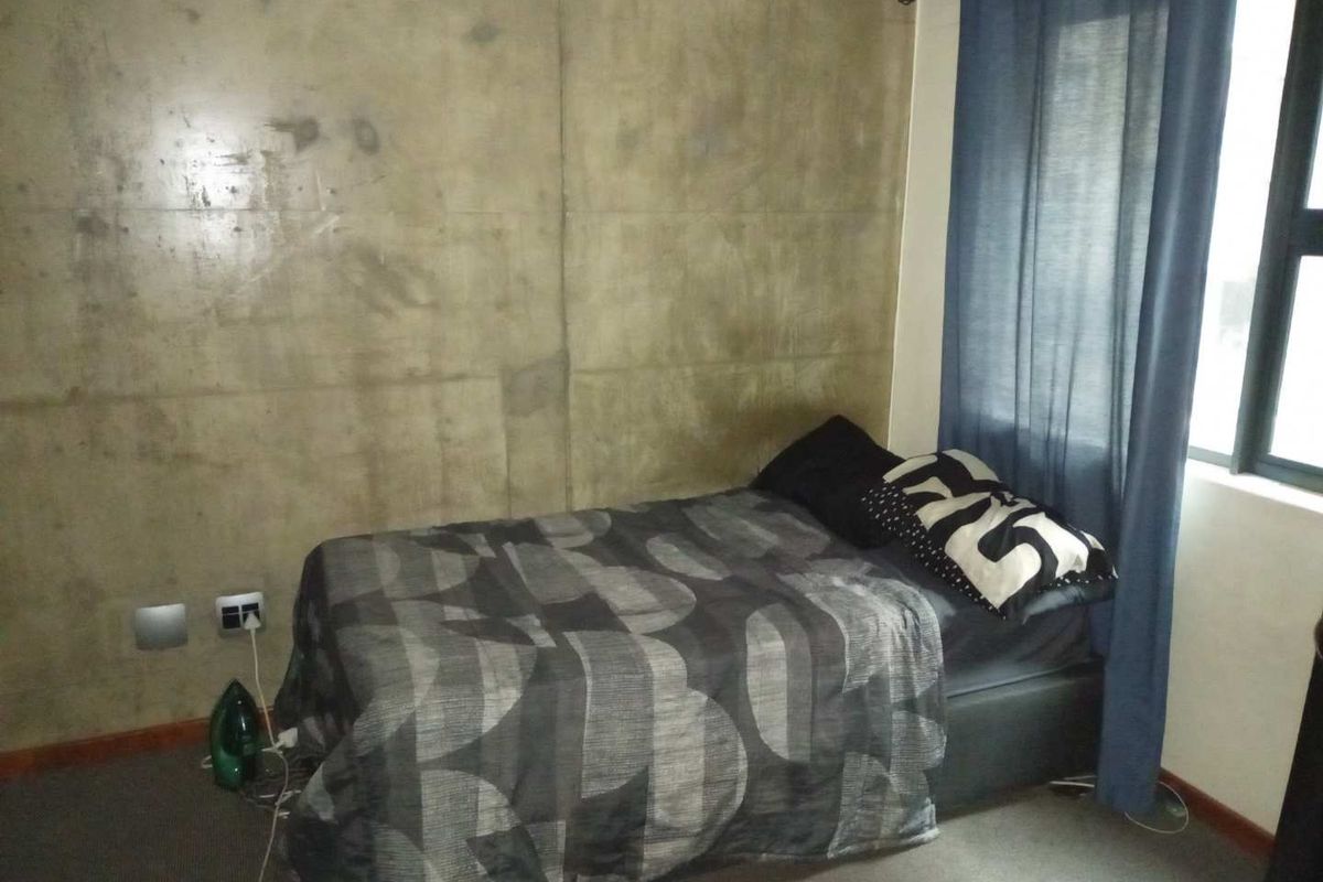 A bedroom featuring an exposed concrete feature wall, carpeted flooring, and a window with curtain rail.