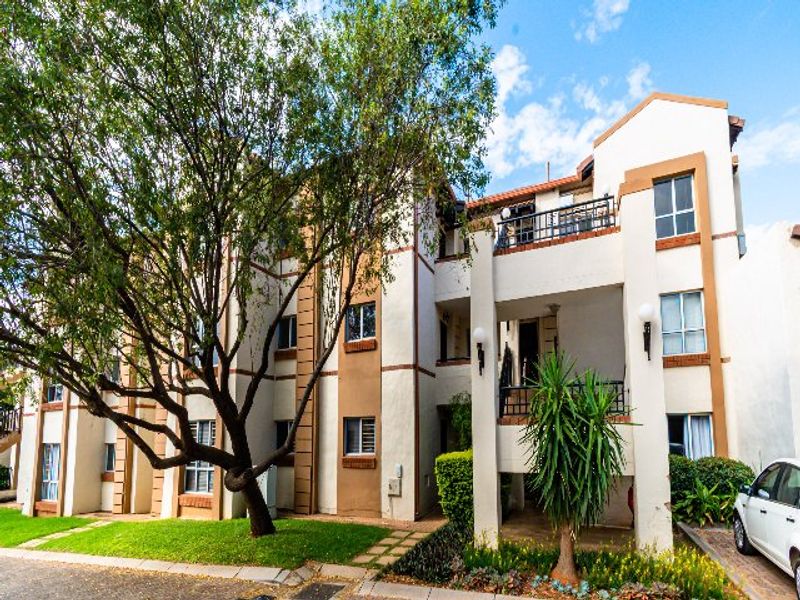 A multi-story apartment building facade featuring balconies with wrought iron railings, neutral exterior paint, and a paved driveway area.