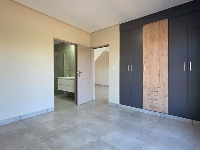 A modern, empty bedroom featuring sleek built-in cupboards with a wood-accent finish, neutral tiled flooring, and direct access to an en-suite bathroom.