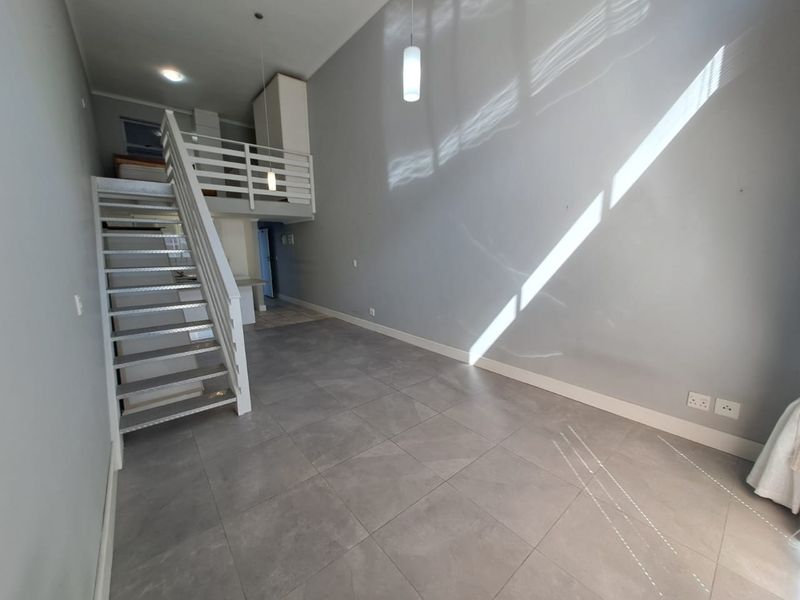 An open-plan living area featuring high ceilings, a mezzanine level with a metal staircase, and modern tiled flooring throughout.