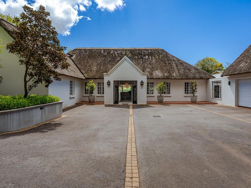 A well-maintained front facade featuring a traditional thatched roof, paved driveway, and a secure entrance with wrought iron gates.