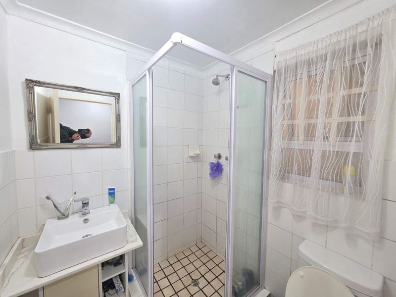 A functional bathroom featuring a corner shower enclosure, a modern basin, and a toilet, with white wall tiling and a window for natural light.