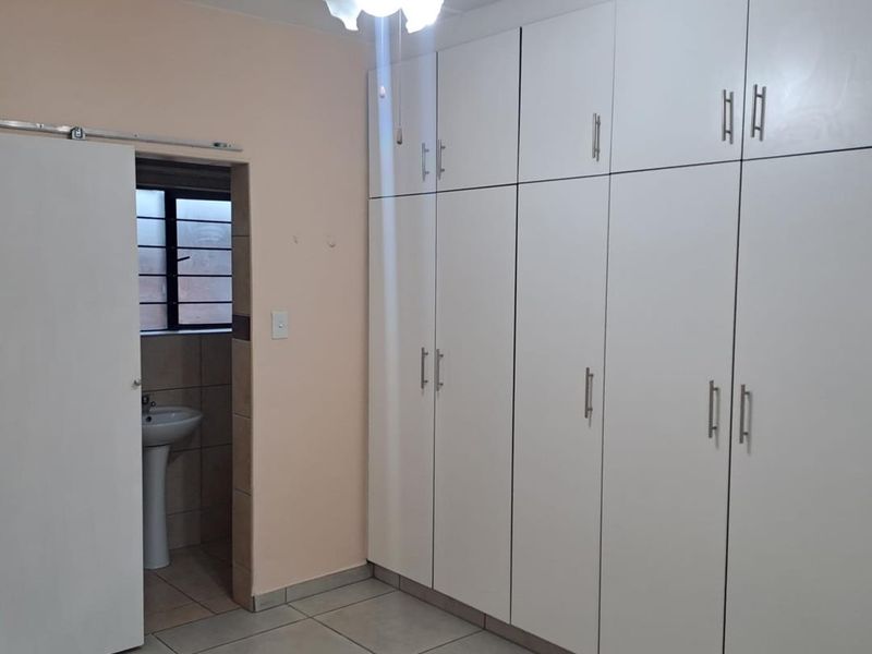A clean, empty bedroom featuring extensive built-in white cabinetry, tiled flooring, a ceiling fan, and direct access to an en-suite bathroom with a pedestal basin.
