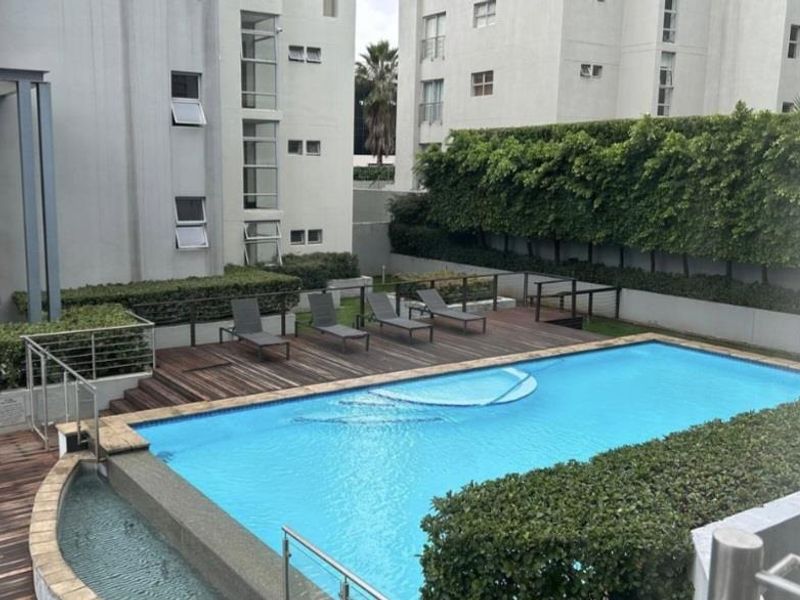A well-maintained communal swimming pool area featuring timber decking, stainless steel safety railings, and manicured garden hedging within an apartment complex.