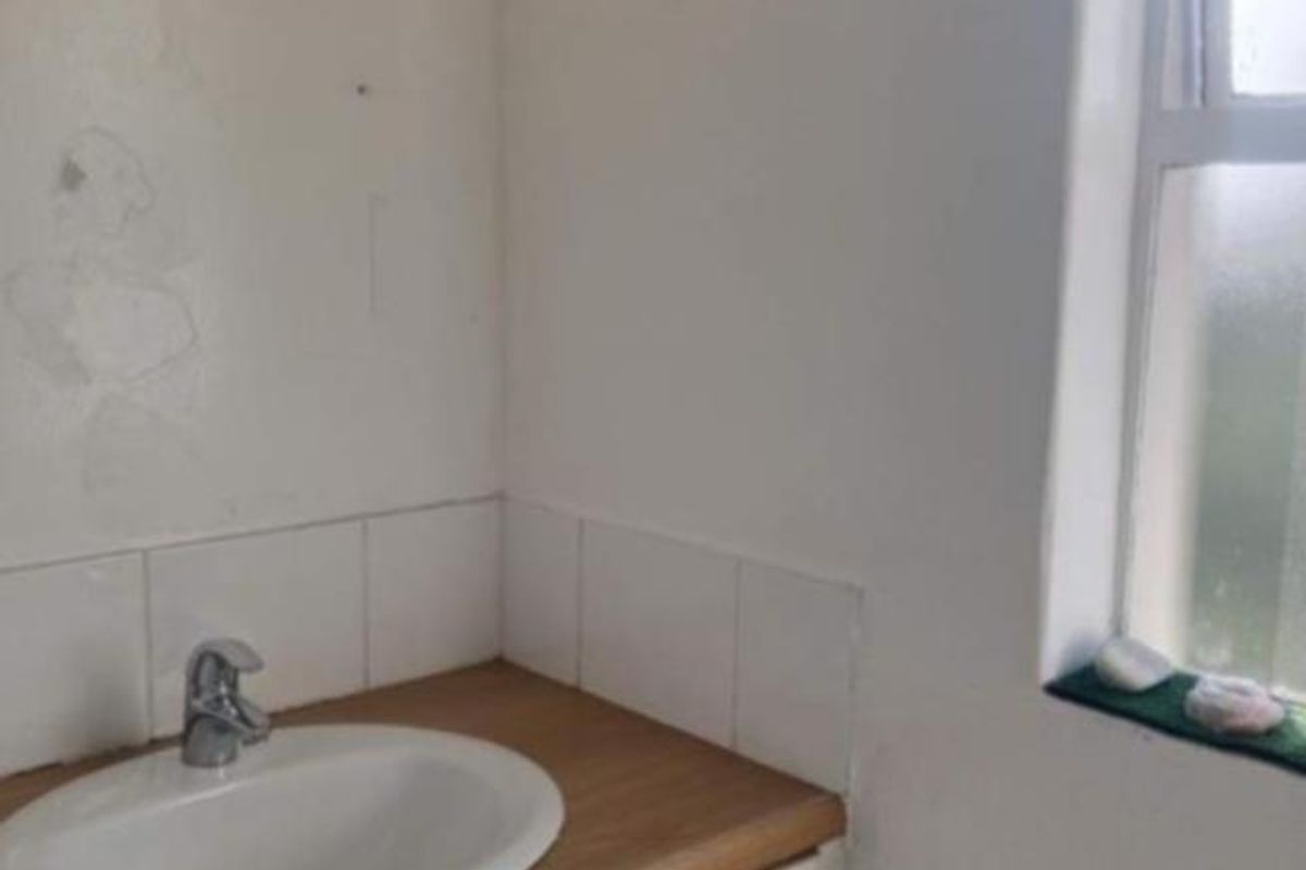 A compact bathroom featuring a vanity cabinet with a basin, tiled splashback, and wall-mounted storage shelf, requiring cosmetic updates.