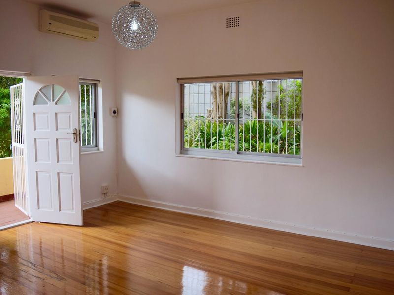 A bright, empty bedroom featuring polished hardwood flooring, a wall-mounted air conditioning unit, and security bars on the windows.