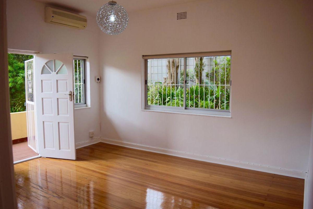 A bright, empty bedroom featuring polished hardwood flooring, a wall-mounted air conditioning unit, and security bars on the windows.