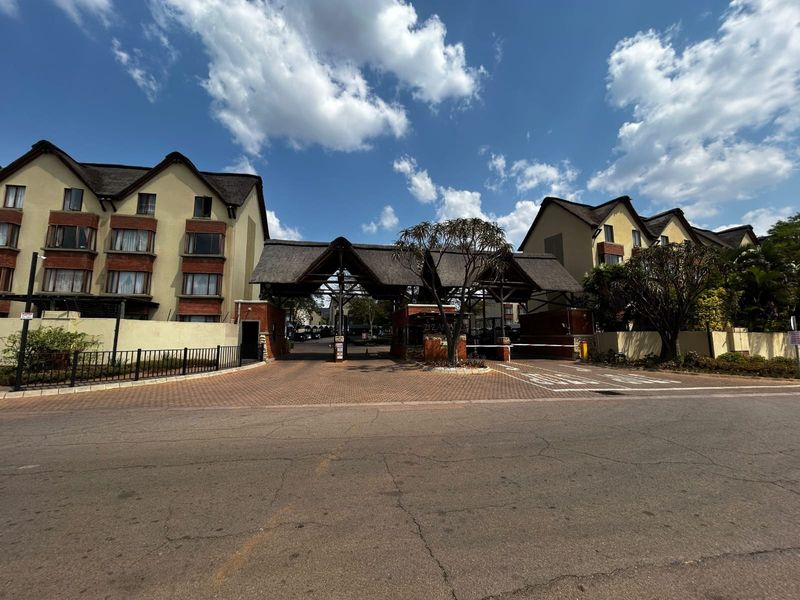 The main entrance to the apartment complex features a secure gatehouse with a thatched roof, brick detailing, and a paved driveway.