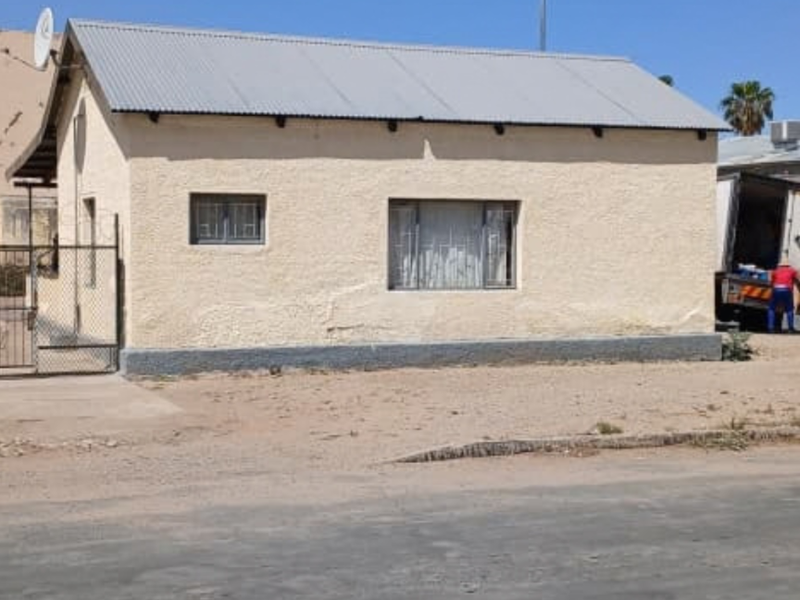 Street-facing view of a single-story commercial building featuring a corrugated metal roof, plastered exterior walls, and security bars on windows.