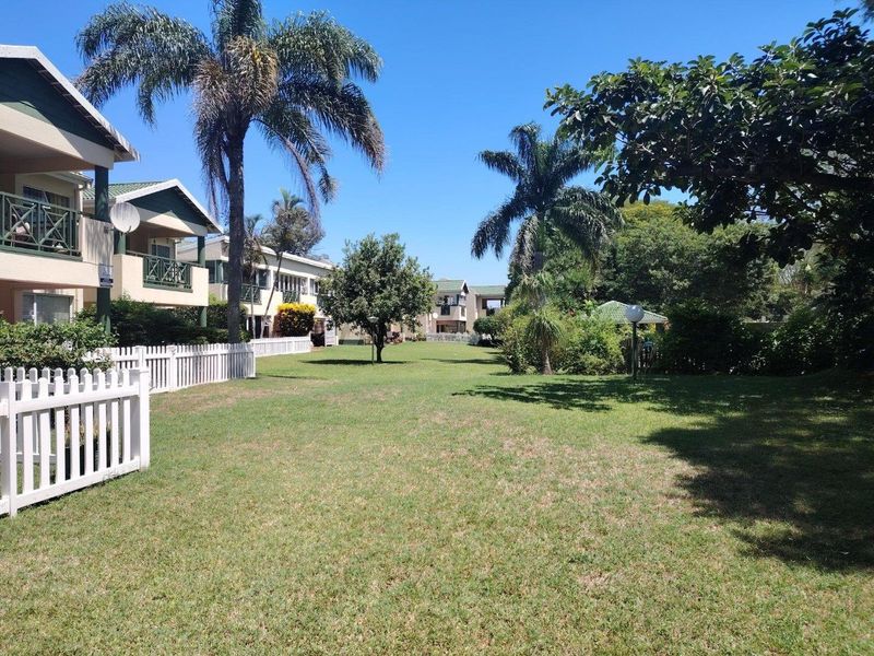 A well-maintained communal garden area featuring a manicured lawn, mature palm trees, and white picket fencing surrounding the apartment complex.