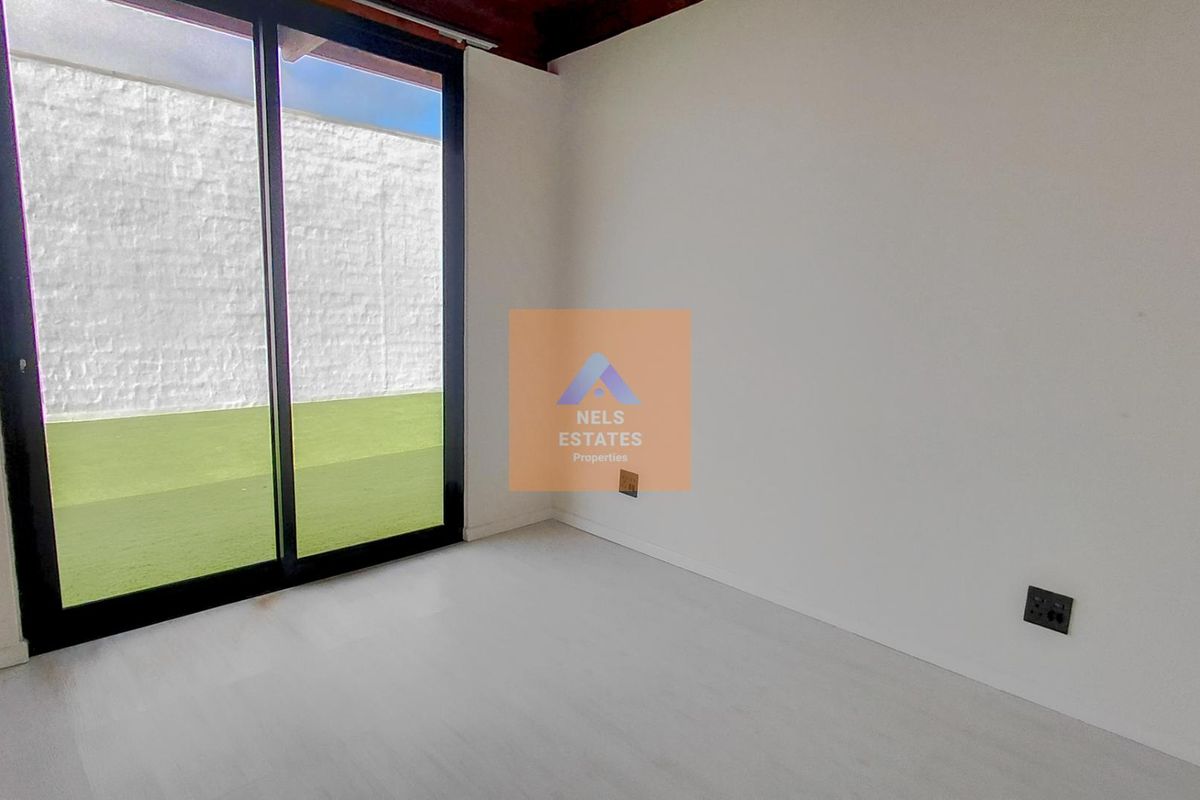 A bright, empty bedroom featuring modern light-toned flooring, exposed timber ceiling beams, and a large sliding glass door providing access to an outdoor area.