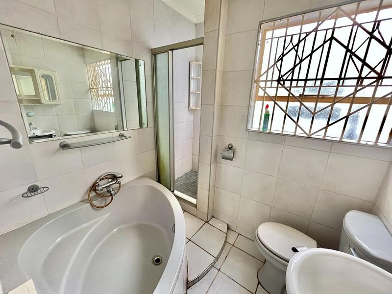 A dated bathroom featuring a built-in bathtub, separate shower enclosure, toilet, and pedestal basin with tiled finishes and security bars on the window.