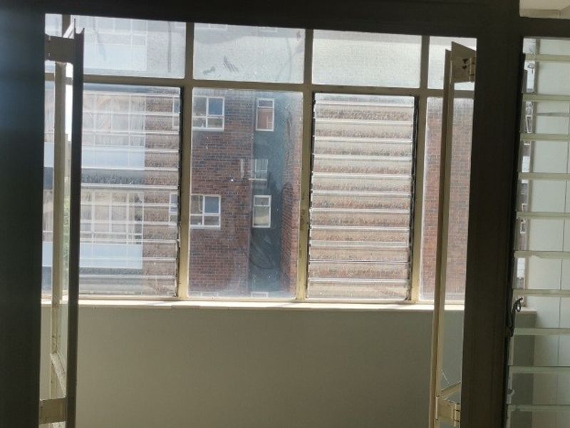 An enclosed balcony area featuring metal-framed louvre windows and security bars, offering a view of the adjacent apartment building.