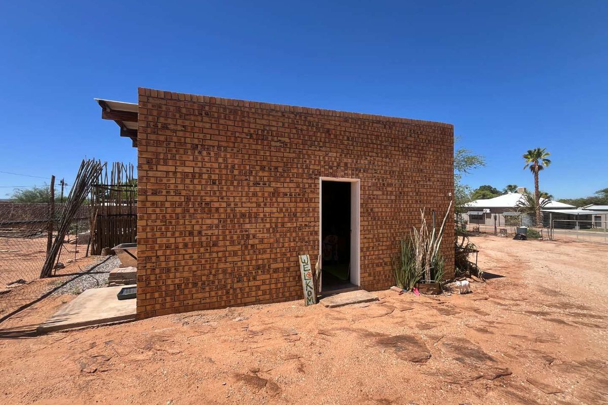 A detached brick structure featuring a simple entrance, set on an unpaved, sandy plot with basic perimeter fencing.