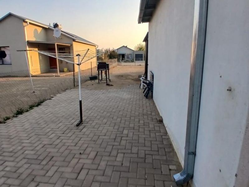 A rear exterior view showing a paved patio area alongside the house wall with a metal downpipe and surrounding fencing.